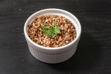 Cooked buckwheat porridge in a deep plate with a leaf of parsley on a black wooden background. Top view