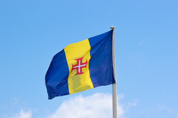 Waving flag of Madeira with blue sky in the background. It consists of a blue-gold-blue vertical triband with a red-bordered white Cross of Christ in the center. Portuguese island in Atlantic ocean