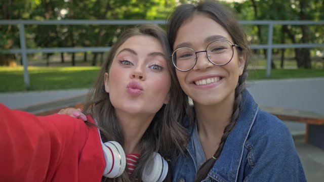 Cheerful beautiful young girls friends gesturing with hands and grimacing while making selfie photo sitting in the park