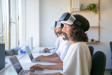 Excited users testing VR simulator. Man and women wearing VR headset, sitting at workplaces with laptops, turning face up. Virtual reality simulation concept