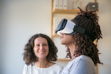 Closeup of young woman in VR headset enjoying experience. Her female colleague looking at her and...