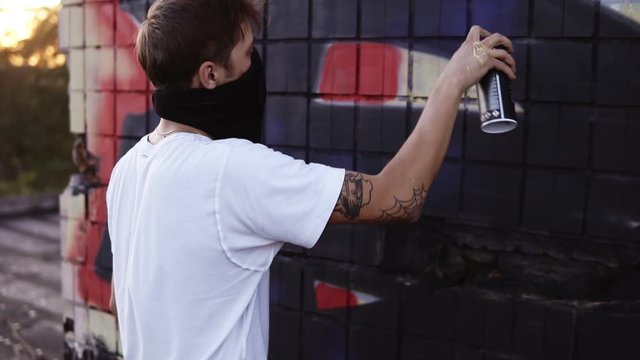 Footage of a young guy in a white T shirt and black scarf on a face standing next to a wall with a graffiti and continuing to paint the picture with a black coloured spray. Backside view. Outdoors
