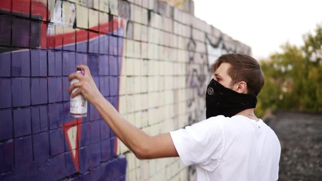 Side view of a young caucasian guy in white T shirt and black scarf on a face applying the blue paint using an aerosol on a building surface outdoors. Hobby, vandalism, art concept