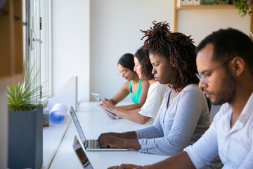 Team of colleagues working on project. Line of office employees sitting at long desk, using laptops and tablets. Working in office concept