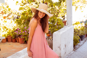 Beautiful brunette in straw hat and long pink dress admires alleys of little white town.