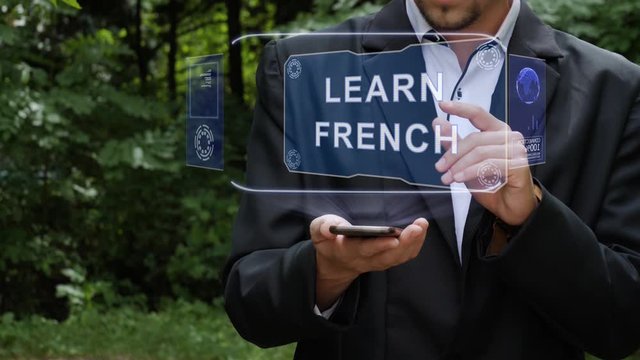 Unrecognizable businessman activates conceptual HUD holograms on smartphone with text Learn French. Bearded man in a white shirt and a jacket with a holographic screen on a background of green trees