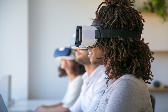 Excited African American user enjoying VR experience. Diverse group of people wearing VR headset, sitting at long desk. Virtual reality experience concept