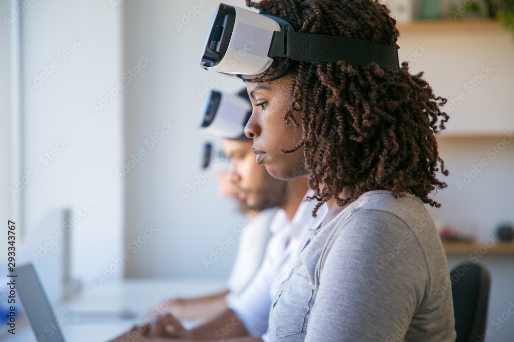 Wall mural Serious African American VR designer working in office. Young black woman wearing VR headset, sitting at long desk with colleagues. Virtual reality developer concept