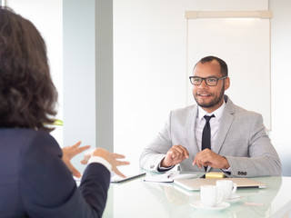 Fototapeta na wymiar Smiling businessman talking with colleague. Young African American businessman in eyeglasses sitting at table with female colleague during business meeting. Job interview concept