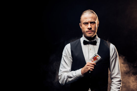 handsome bearded croupier holding playing cards on black with smoke