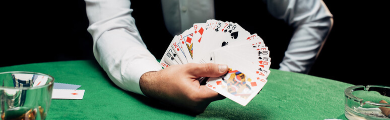 panoramic shot of croupier in formal wear holding playing cards near poker table isolated on black