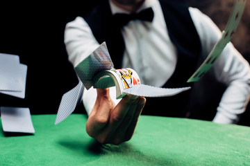 cropped view of croupier throwing in air playing cards near poker table on black
