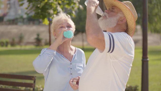 Middle-aged caucasian couple drinking tasty water in the summer park and chatting. Positive mature man in straw hat spending time with his adorable wife outdoors. Active life after retirement.
