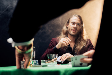 selective focus of handsome bearded man in eye glasses playing poker with friend on black with smoke