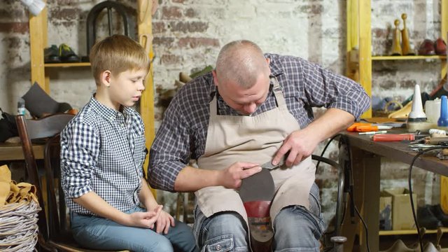 Zoom in of cheerful middle aged shoemaker repairing shoe sole and explaining process to little son sitting beside him in workshop