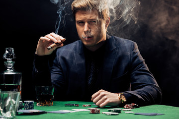 handsome man smoking cigar near poker table on black with smoke
