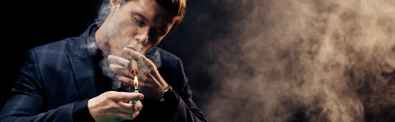 panoramic shot of handsome man holding lighter while smoking on black with smoke