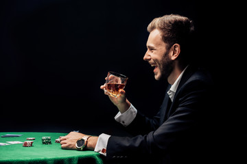 side view of happy bearded man smiling and holding glass near poker table isolated on black