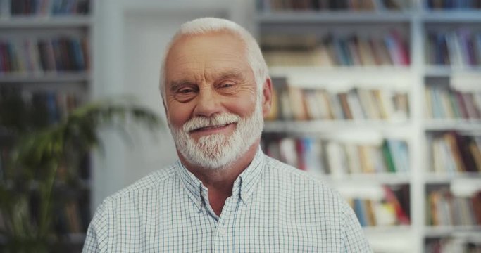 Portrait of the handsome senior Caucasian man with a grey hair and beard looking staright to the camera and smiling in the library. Close up.