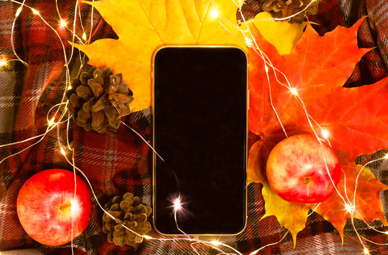 View from above. Mobile phone or smartphone, touch screen. flat lay. Autumn mood, plaid cage, apples and cones. Cozy Homely atmosphere.
