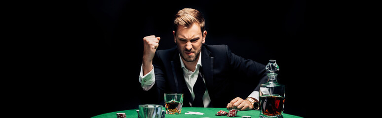 panoramic shot of angry man with clenched hand near alcohol and playing cards isolated on black