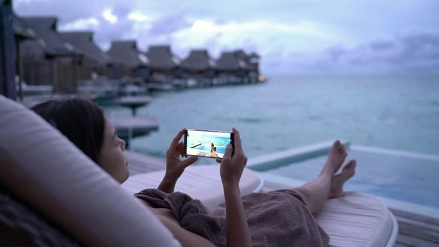 Phone - woman using mobile cell phone social media app on vacation looking at photos posting images on social network by pool at night on travel holidays. Girl using smartphone app looking at screen.