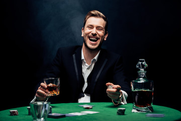 selective focus of cheerful man holding glass with alcohol on black with smoke