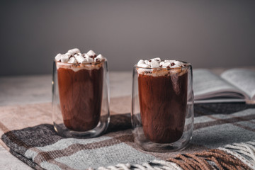 Two glasses of hot cocoa with marshmallows on a brown wool blanket next to an open book