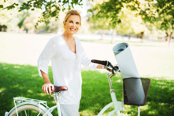 attraktive ältere frau ist mit dem fahrrad im park unterwegs, sie hat eine yoga-Matte dabei 