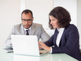 Fototapeta premium Serious coworkers using laptop. Professional multiethnic businessman and businesswoman sitting at table and working with laptop computer in office. Business and technology concept