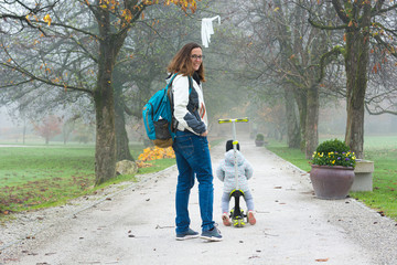Youg mother walking with her daughter riding a toddler scooter in park.