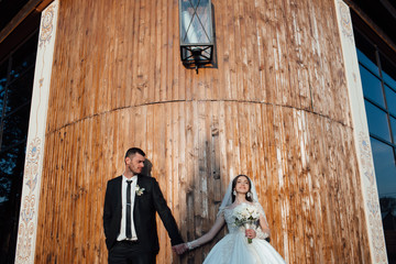 Wedding. The groom in a suit and the bride in a white dress standing  and holding bouquets of white flowers on background of wooden wall. Young and stylish lovers couple. Wedding couple