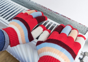 A child in bright warm wool gloves warms his hands on a heating radiator. The symbolic image of the heating season at home. Part of body, selective focus.
