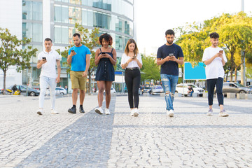 Obraz premium Multiethnic friends walking together and texting messages on smartphones outside. Diverse men and women going down city street and using mobile phones. Communication concept