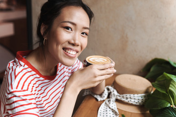 Portrait of joyful asian woman drinking cup of coffee in cafe indoors