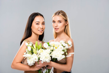smiling blonde and asian brunette naked women with flowers isolated on grey