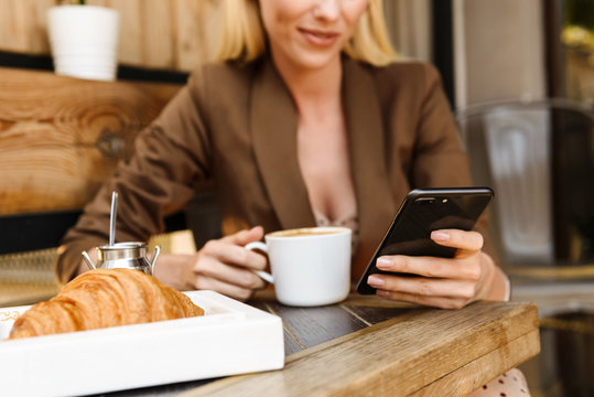 Cropped image of pleased cute woman using cellphone and drinking coffee