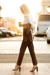 Image of blonde elegant woman drinking coffee takeaway while walking