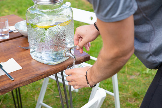 Man pouring lemonade in glass. Close-up partial view of young man holding glass and pouring healthy lemonade from glass jar in backyard. Refreshing beverage concept