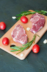 Raw juicy pork steaks with red tomatoes, rosemary and garlic on cutting board, close-up, top view
