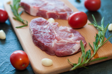 Raw juicy pork steaks with red tomatoes, rosemary and garlic on a cutting board.