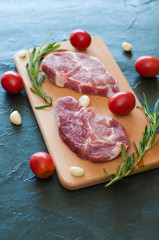 Raw juicy pork steaks with red tomatoes, rosemary and garlic on cutting board, close-up, top view