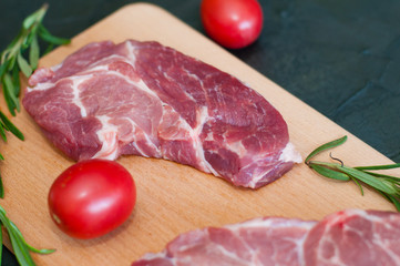Raw pieces of pork, rosemary and tomatoes on a cutting board, on dark gray concrete background, close-up