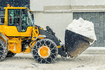 Big loader machine with steel metal chains removing big snow pile from city street at alpine...