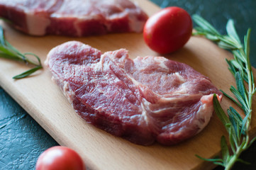 Raw pork steak, rosemary and tomato on a cutting board, on a dark gray concrete background, close up, in dark tonality