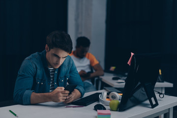 selective focus of young programmer using smartphone while sitting near african american colleague at night in office