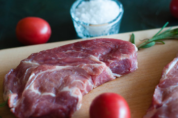 Raw pork steak, salt, rosemary and tomato on cutting board, on a dark gray concrete background, close up, in dark tonality