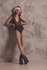 Naklejka premium Fashionable confident woman with long legs in black sexy bodysuit, posing in studio with beton background. Girl in santa clous hat