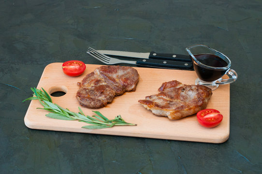 Grilled meat steaks of pork, veal or beef, with rosemary, soy sauce and tomato on cutting board, fork and knife