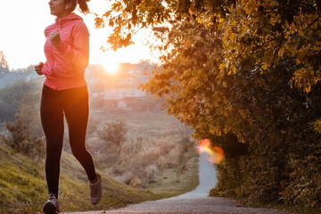 Girl runner running on sunset in autumn forest and park wearing pink jacket. Young woman jogging in fall colors. Sport and activities concept.